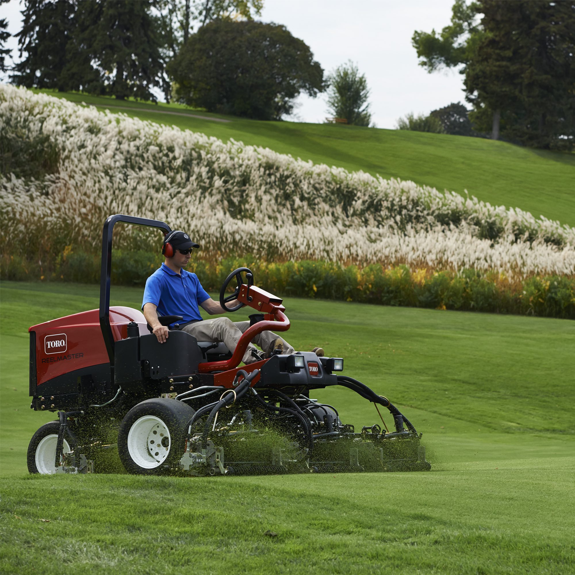 A person operates a Toro mower on a lush green field with tall grass and trees in the background