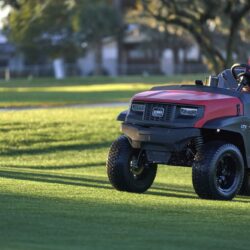 A man drives a red Toro utility vehicle across a green lawn in a park setting