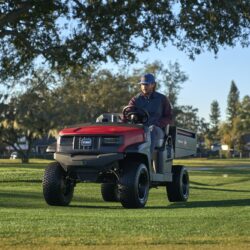 A man drives a red utility vehicle across a grassy area under a clear sky