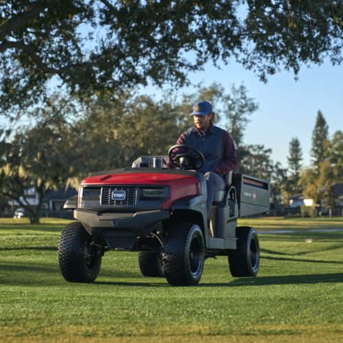 A man drives a red utility vehicle across a grassy area under a clear sky
