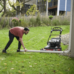 A man adjusts a Toro lawn mower on a grassy yard near a house and garden