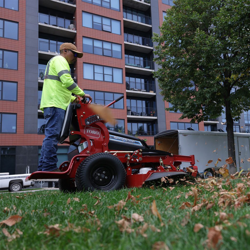 A user operates a red stand-on mower on grass, surrounded by urban buildings and autumn leaves