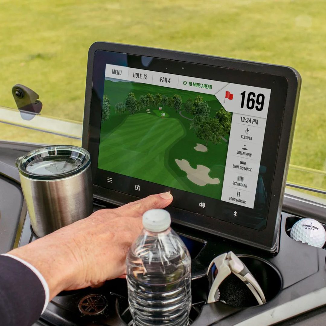 Yamatrack Side View Golfer Close-up of a touchscreen display showing golf course graphics with a hand interacting, beverage, and golf balls nearby