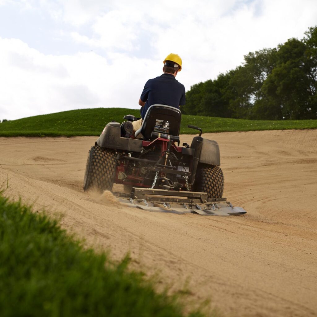 A person operates a Toro Sand Pro 3040, raking sand on a golf course bunker