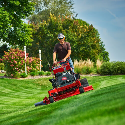 assetstorage-1 A man operates a red stand-on mower on a grassy slope in a landscaped area