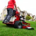 A person operates a red Toro stand-on mower on a grassy lawn near residential homes
