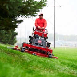 A person operates a red Toro stand-on mower on a grassy slope near trees