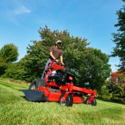 A person operates a red stand-on mower on green grass under a clear blue sky