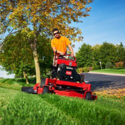 A person operates a red Toro mower on a grassy area with trees and blue sky in the background