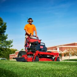 A person in an orange shirt operates a red Toro stand-on mower on green grass