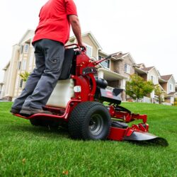 A person operates a red Toro stand-on mower on a grassy lawn near residential homes
