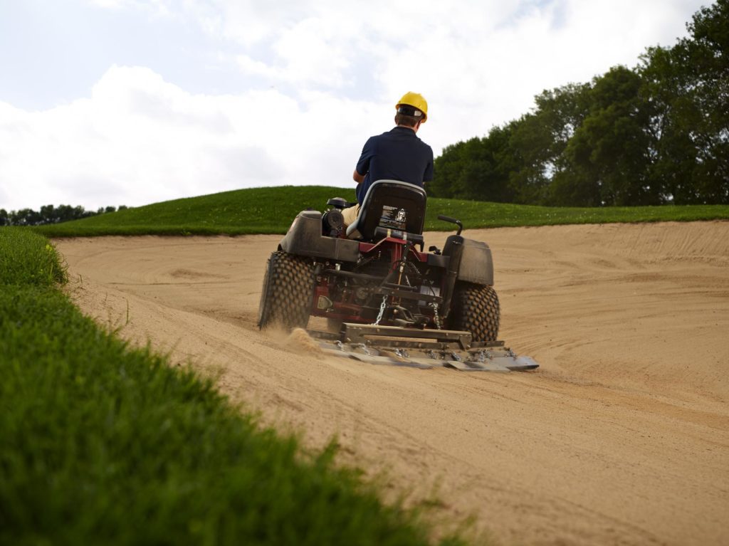 A person operates a sand rake on a golf course bunker, showcasing its maneuverability on sand