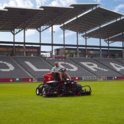 Red ride-on mower in action on a well-maintained grass field with stadium seating in the background