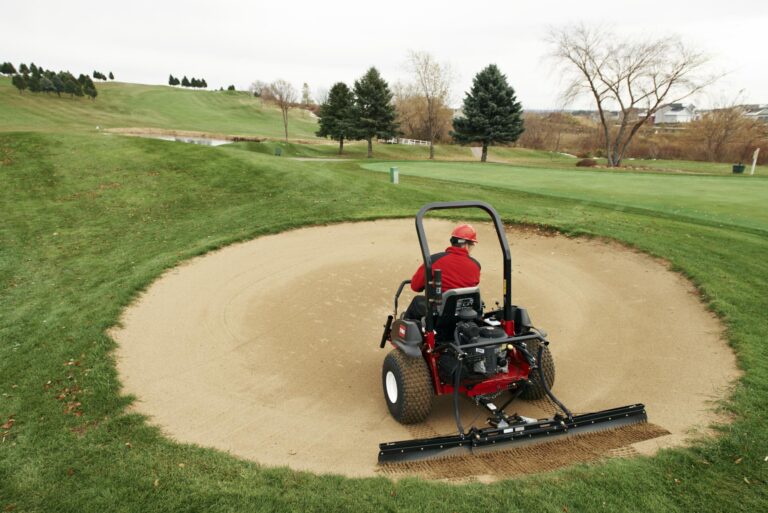 A person operates a sand rake on a golf course bunker, maintaining the sand surface