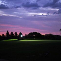 A silhouette of an electric greens roller operates on a golf course at dusk, illuminated by its headlights