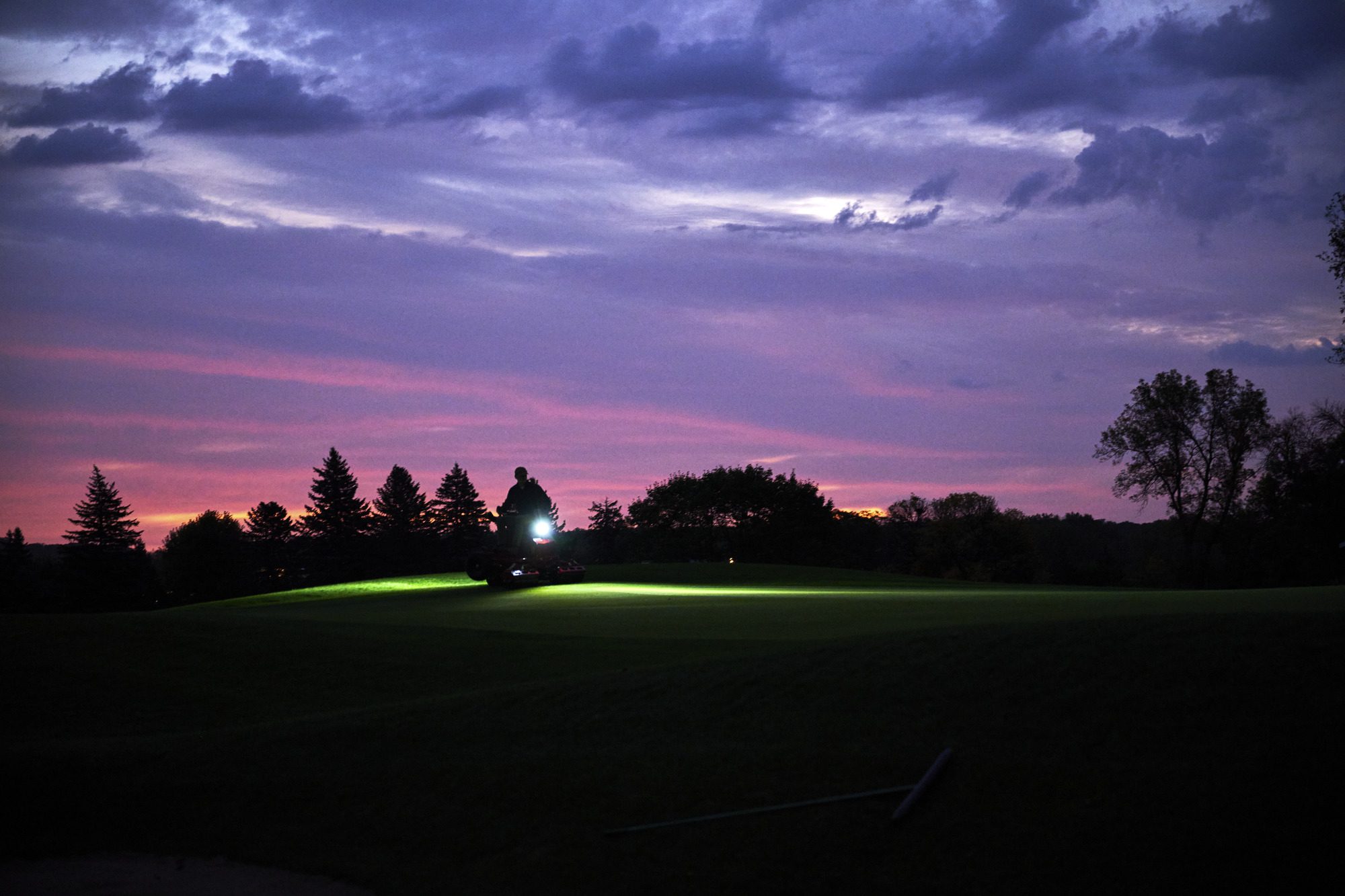cm25_6197L_GreensPro_e1700_010 A silhouette of an electric greens roller operates on a golf course at dusk, illuminated by its headlights