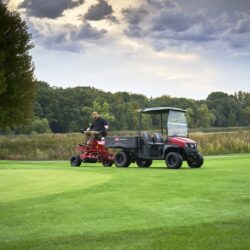 A person operates an electric greens roller on a lush green golf course under a cloudy sky