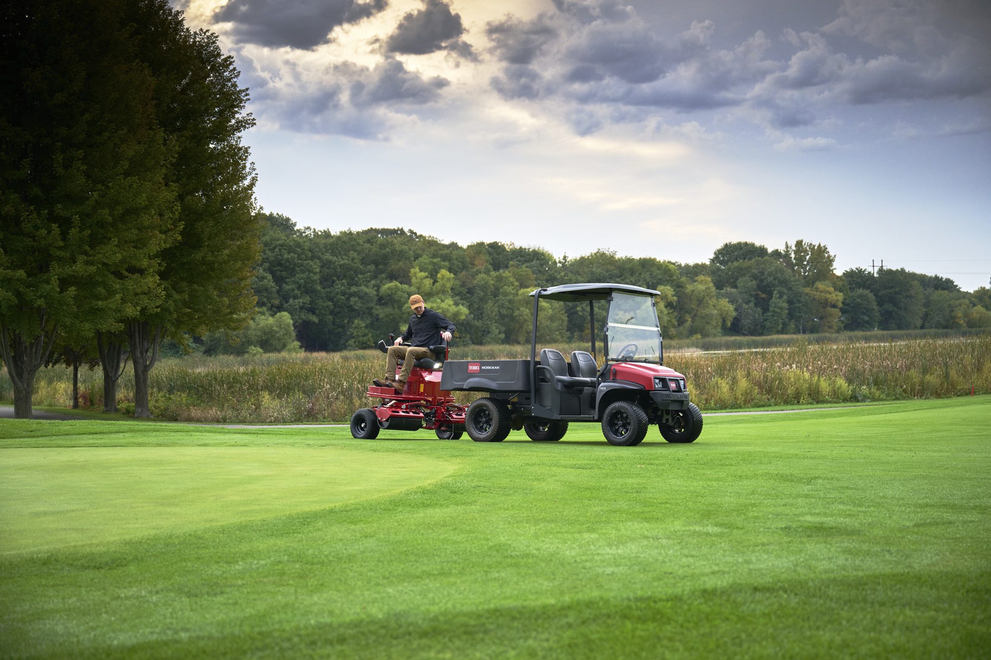 cm25_6197L_GreensPro_e1700_084 A person operates an electric greens roller on a lush green golf course under a cloudy sky