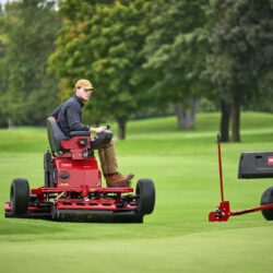 A person operates a red electric greens roller on a lush green golf course