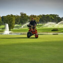 A person operates a red electric greens roller on a golf course with fountains in the background