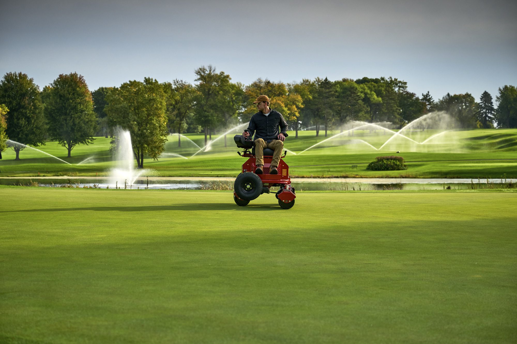cm25_6197L_GreensPro_e1700_152 A person operates a red electric greens roller on a golf course with fountains in the background