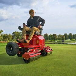 A man operates a red electric greens roller on a golf course, surrounded by trees and a fountain