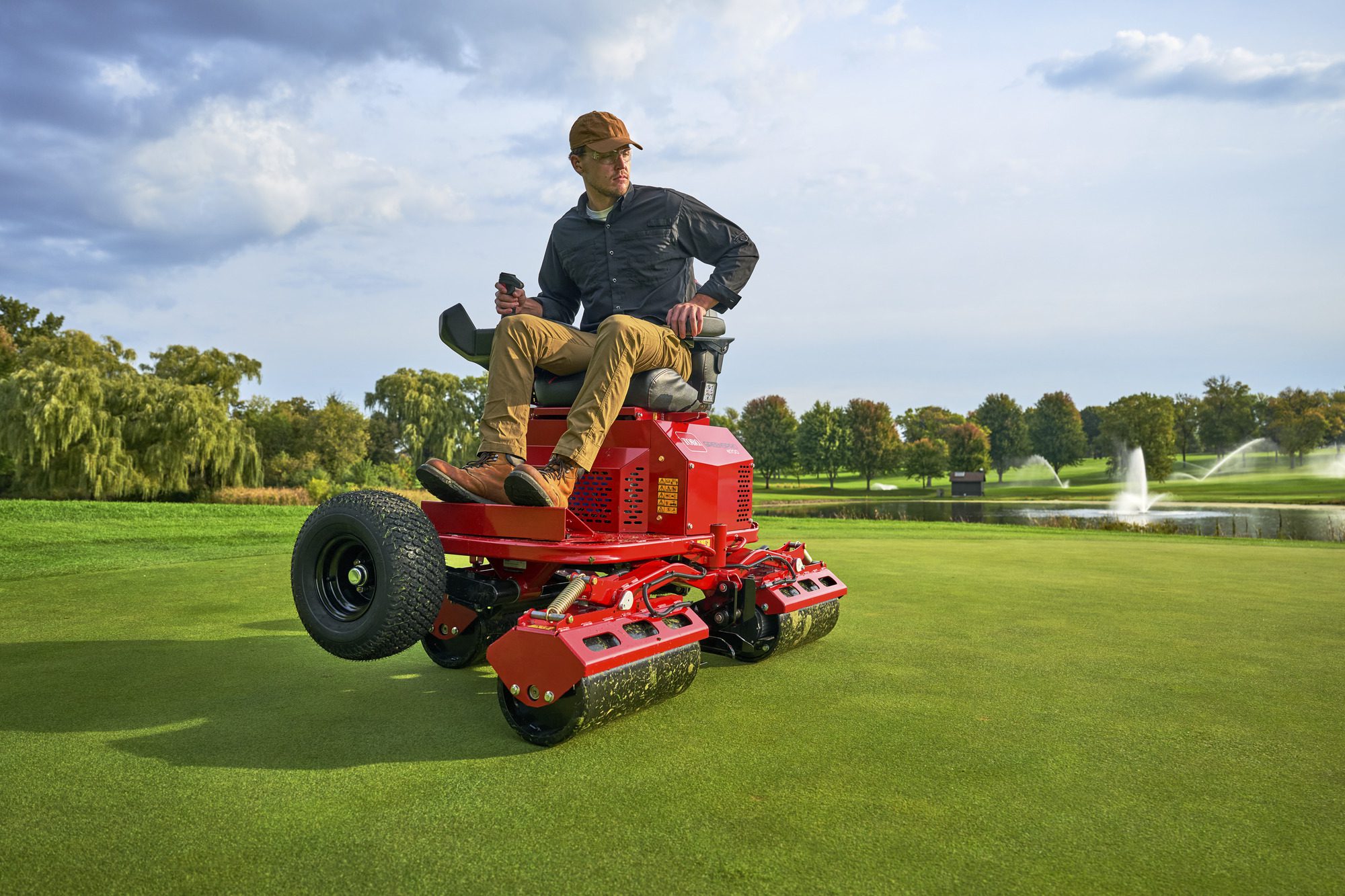 cm25_6197L_GreensPro_e1700_155 A man operates a red electric greens roller on a golf course, surrounded by trees and a fountain