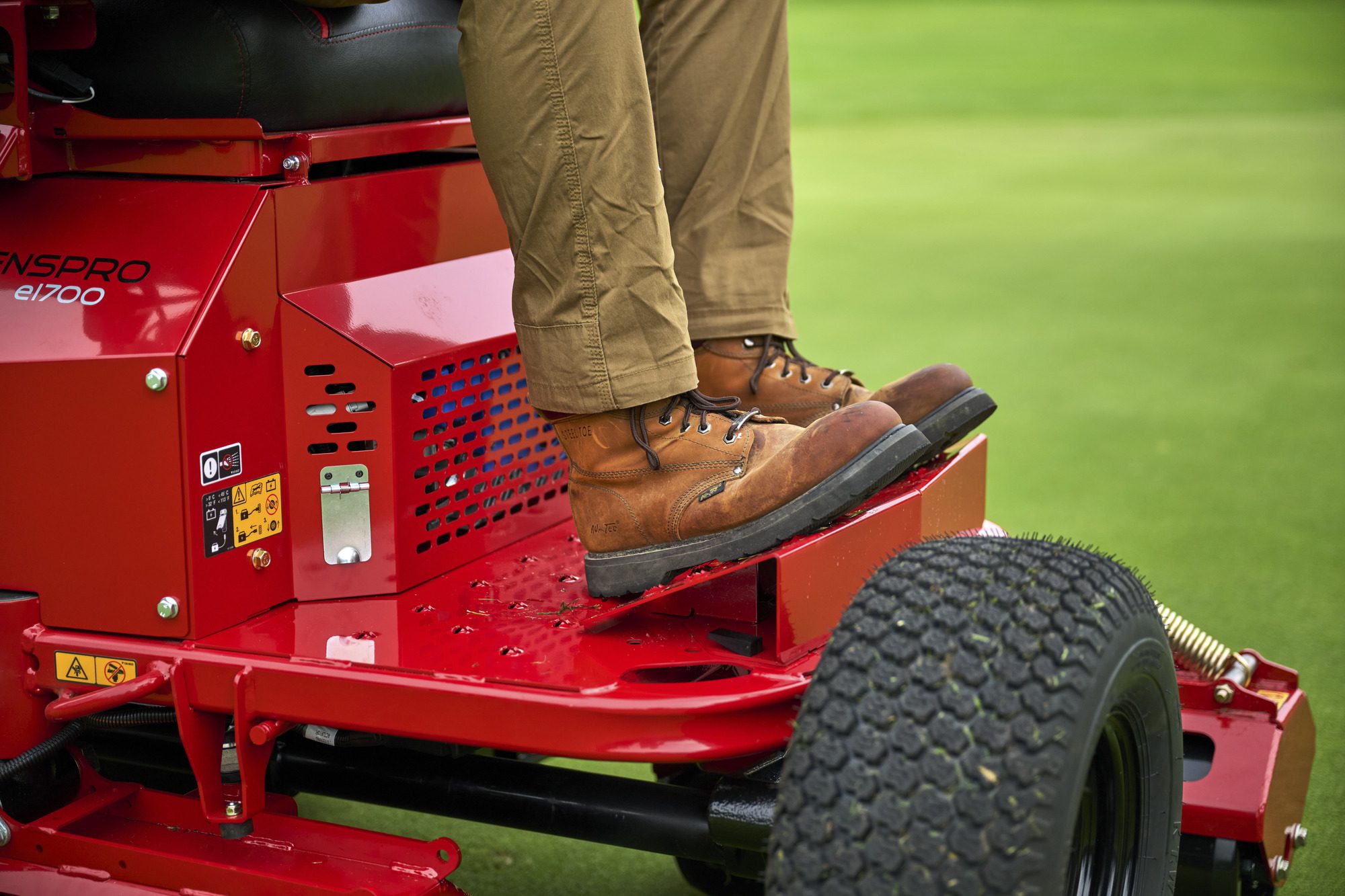 cm25_6197L_GreensPro_e1700_207 Close-up of a person’s feet on the red electric greens roller, showcasing its design and control features