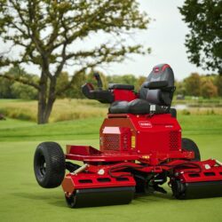 A red electric greens roller with a seat, joystick, and wide rollers on a golf course