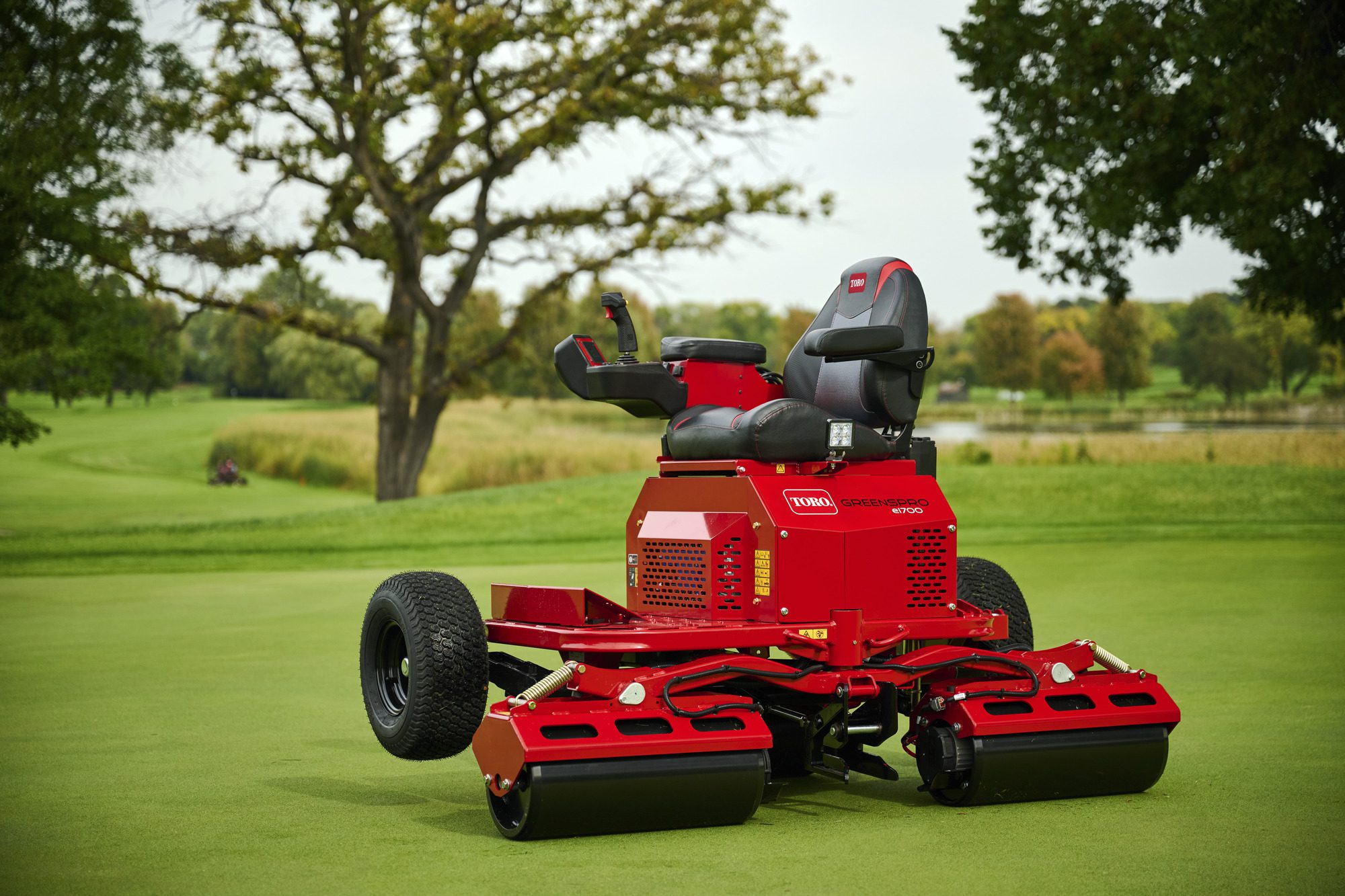 cm25_6197L_GreensPro_e1700_237 A red electric greens roller with a seat, joystick, and wide rollers on a golf course