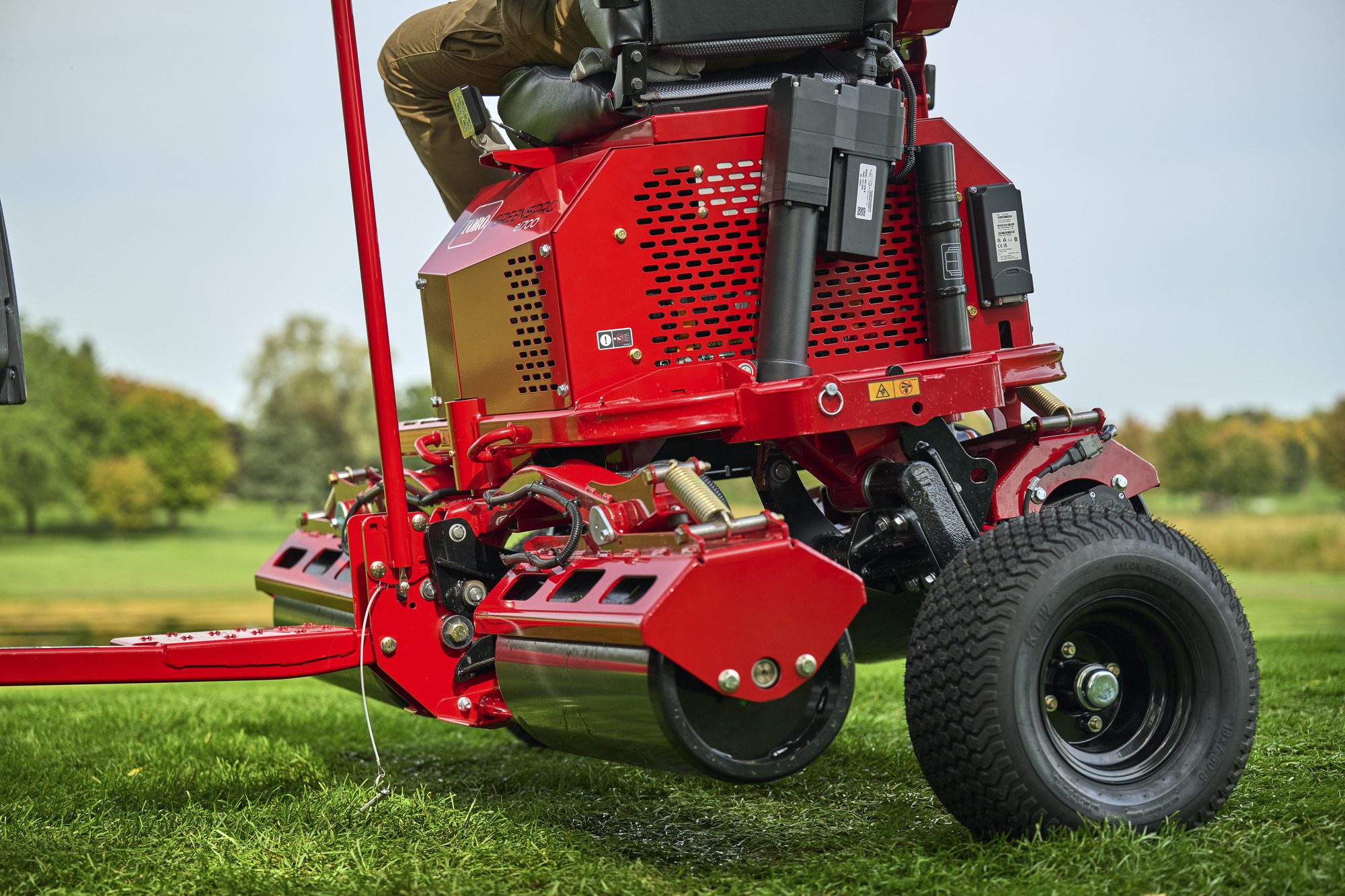 cm25_6197L_GreensPro_e1700_293 Close-up of a red electric greens roller with a rider, showcasing its design and key components on grass