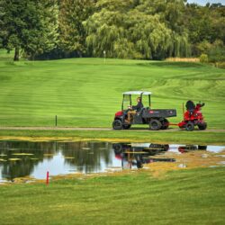 A rider operates an electric greens roller near a pond on a lush golf course