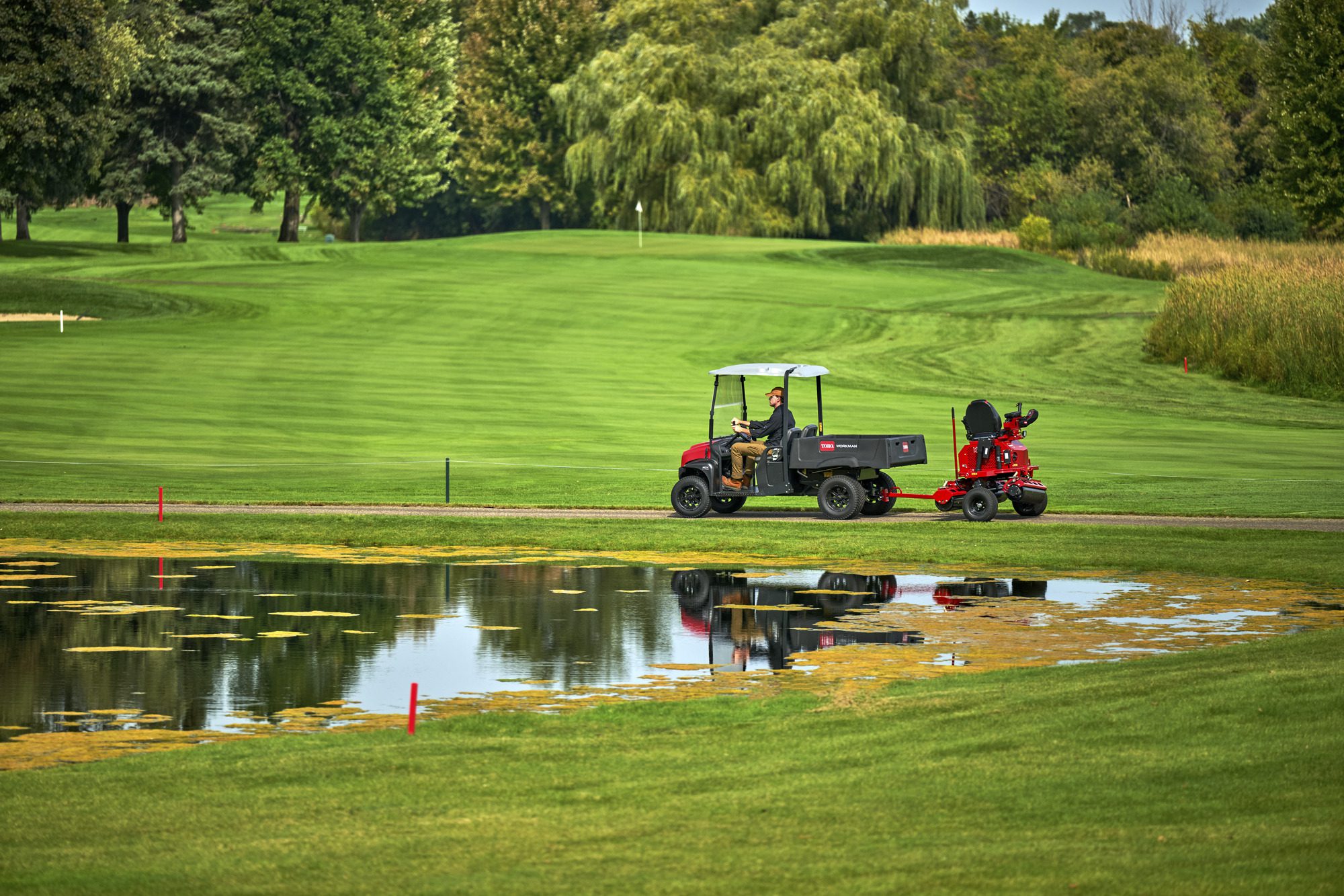 cm25_6197L_GreensPro_e1700_297 A rider operates an electric greens roller near a pond on a lush golf course