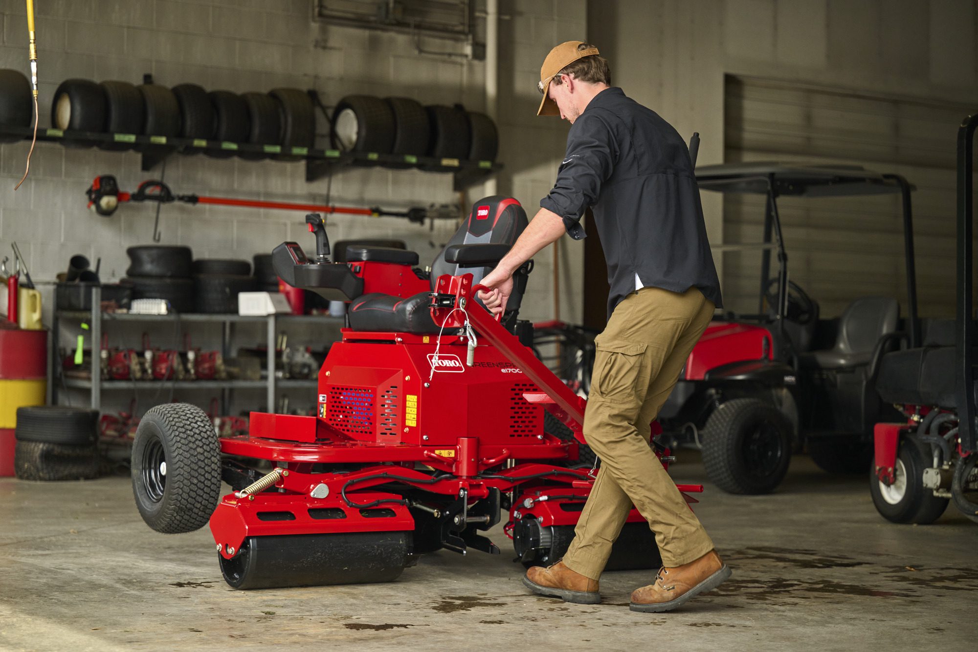 cm25_6197L_GreensPro_e1700_310 A man operates a red electric greens roller in a garage with various equipment in the background
