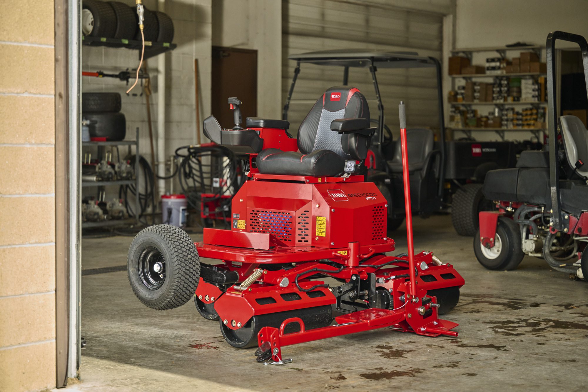 cm25_6197L_GreensPro_e1700_330 A red electric greens roller with joystick controls, parked in a garage with tools and equipment in the background