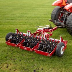 Red tow-behind aerator with multiple tines attached to a tractor on green turf grass