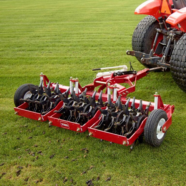 Red tow-behind aerator with multiple tines attached to a tractor on green turf grass