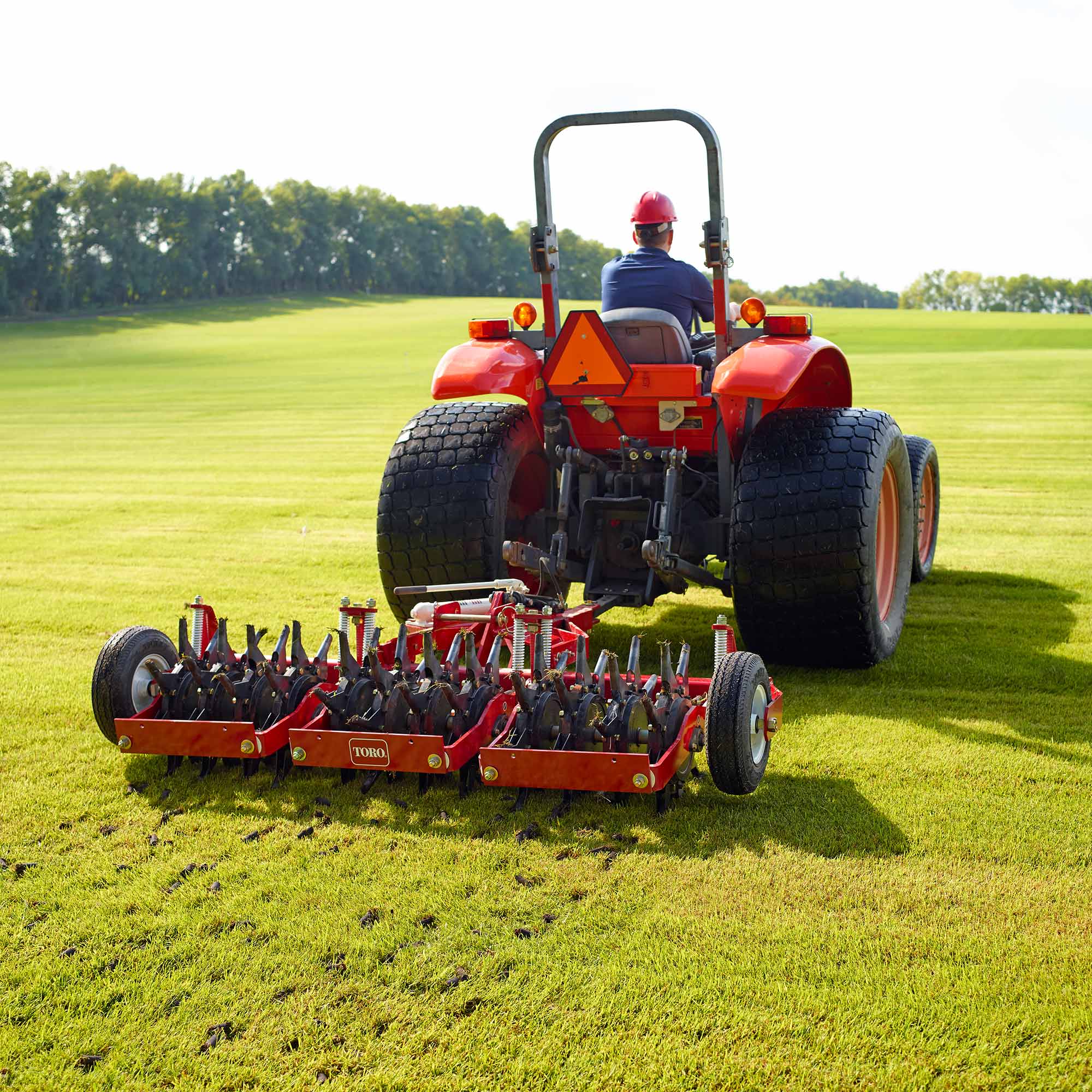 feat4 A person operates a red Toro aerator attached to a tractor, aerating a green turf field