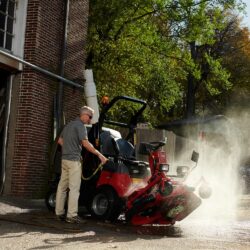 A man cleans a red Toro ride-on mower with water, showcasing its out-front deck and collection system