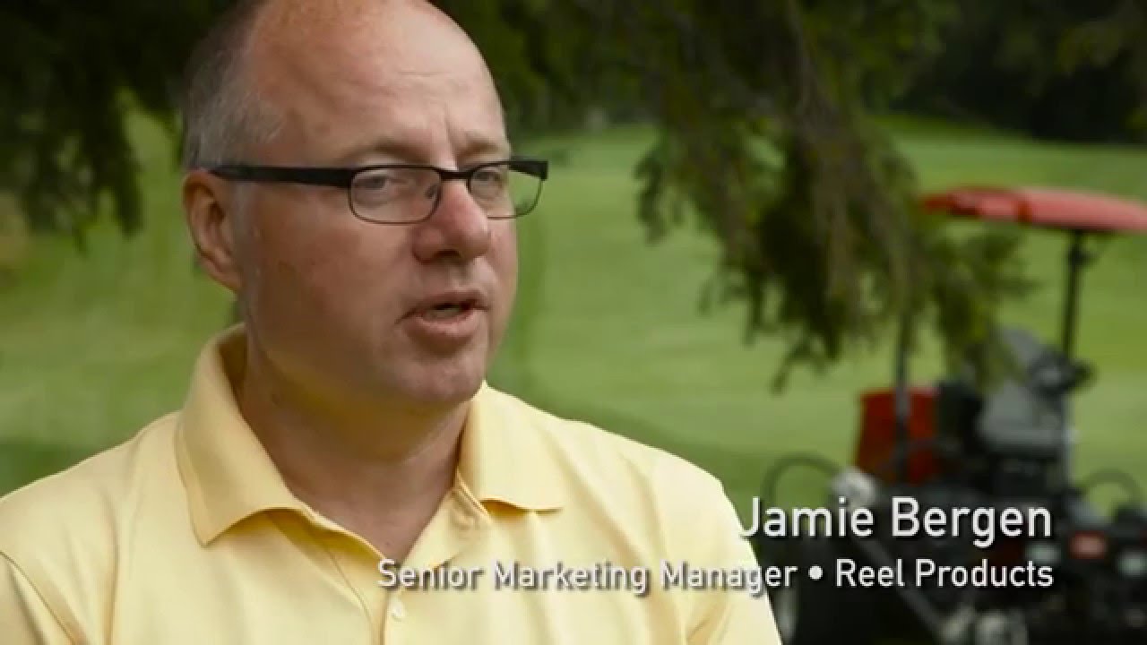 A man in a yellow shirt discusses reel innovations with a mower in the background